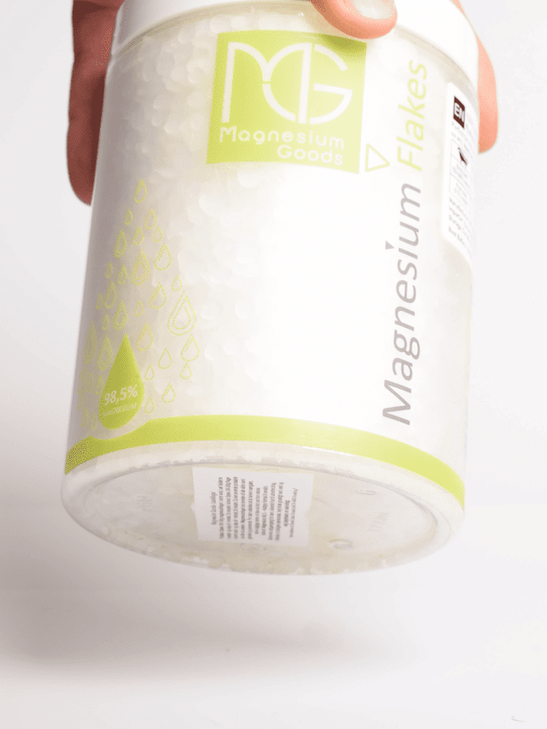 Hånd som holder en hvit krukke med Magnesium Flakes-etikett i grønt fra Magnesium Goods