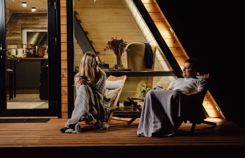 Couple wrapped in blankets chatting on a wooden cabin deck at night, warm interior lights behind them
