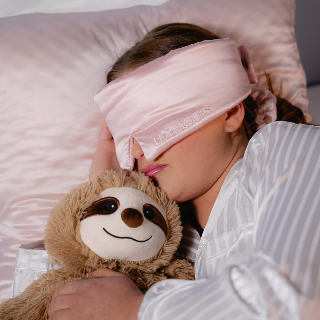 Person sover i stripete pysj med rosa silkesovemaske og klemmer en lodden dovendyrbamse på satengpute