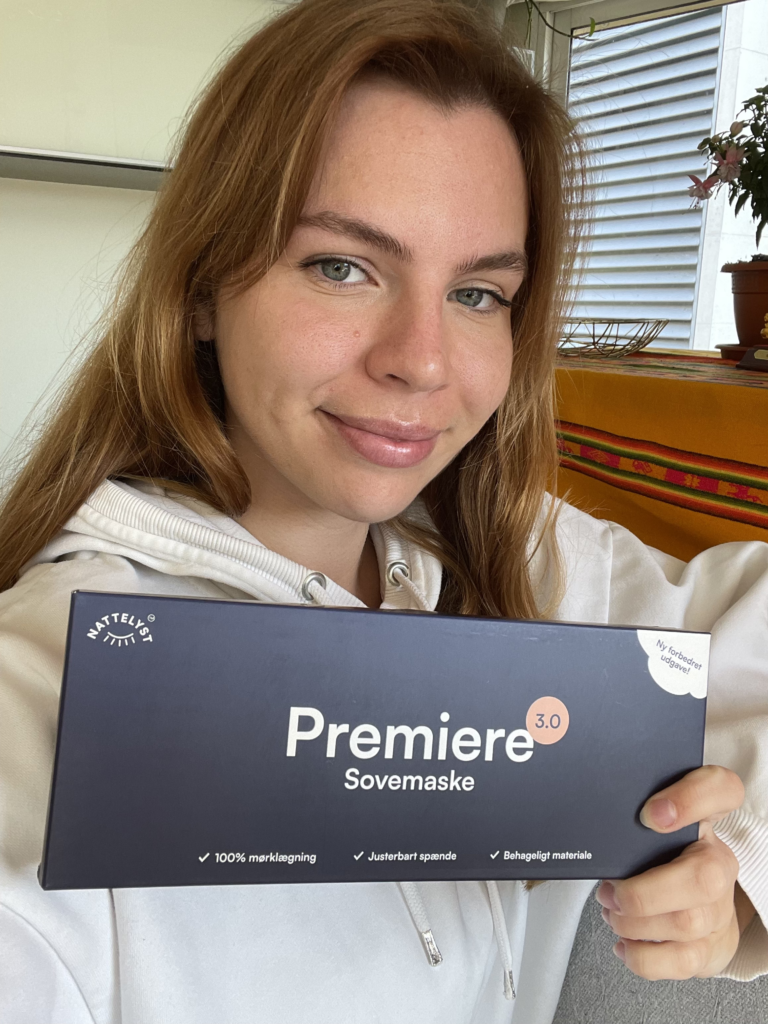 Nærbilde av kvinne med brunt hår som smiler og holder eske med Premiere 3.0 sovemaske