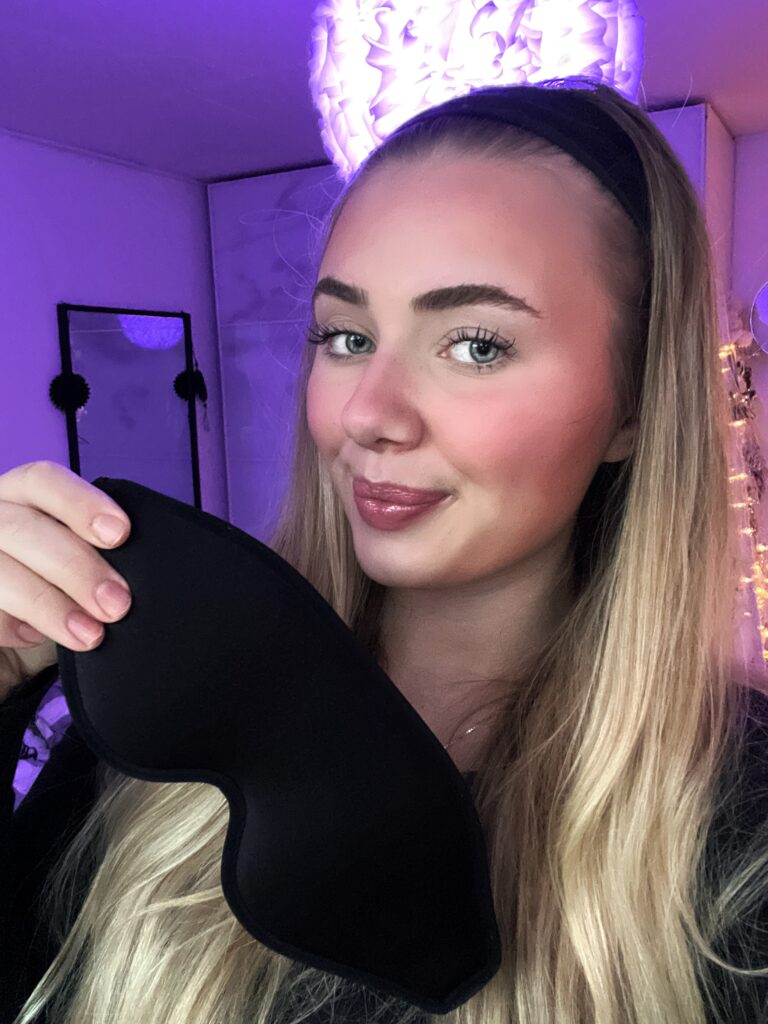 Nærbilde av smilende ung kvinne med langt blondt hår, svart sovemaske og lilla stemningslys