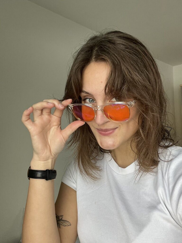 Kvinne med skulderlangt hår i hvit t-skjorte holder oransje speilglassbriller nedover og smiler