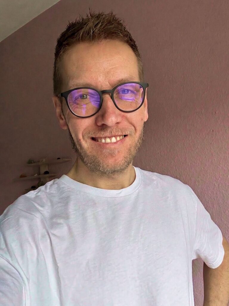 Smilende mann i hvit t-skjorte med runde briller som reflekterer blått lys, foran rosa vegg