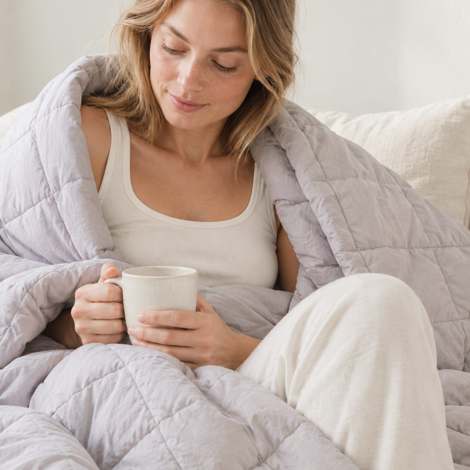 Kvinne pakket i grå vektet dyne holder keramikkopp i fanget, koselig morgenstemning